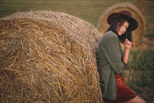 Beautiful stylish woman in hat standing at hay bale in summer evening field. Portrait of young fashionable female relaxing at haystacks, summer vacation in countryside. Tranquility