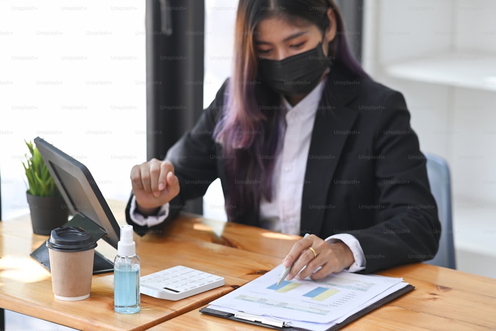 Businesswoman in protective mask working with digital tablet and analyzing financial data on wooden desk.