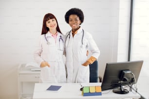 Pretty two young multiethnical women doctors, African and Caucasian, wearing lab coats and stethoscopes, posing embracing each other, standing behind the desk with computer in modern clinic