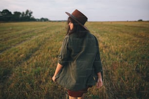 Beautiful carefree woman in hat walking and smiling in evening summer field. Young happy stylish female relaxing in countryside, enjoying evening. Atmospheric moment.