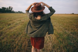 Beautiful carefree woman in hat walking in evening summer field. Young happy stylish female relaxing in countryside, dancing and enjoying evening. Atmospheric moment. Back view