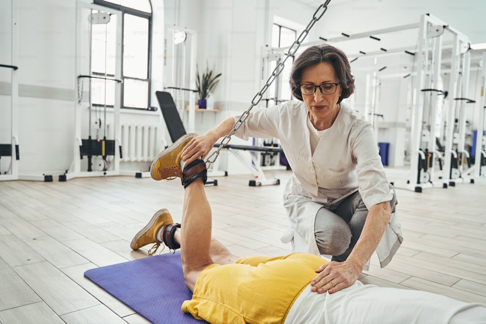 Experienced female physical therapist assisting a Caucasian man with the lying cable leg curl exercise