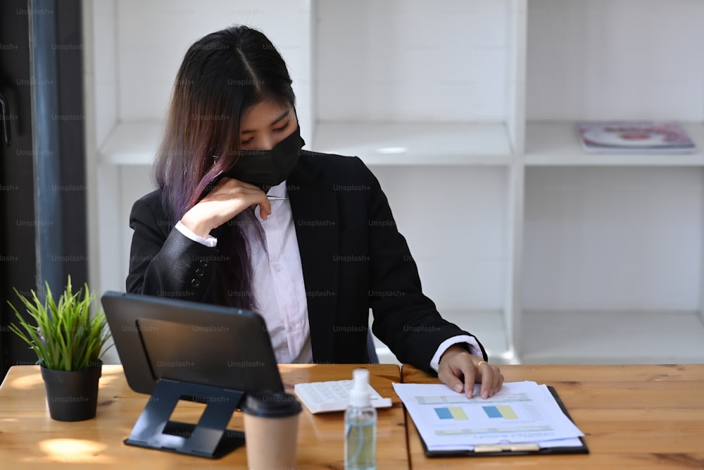 Female accountant in protective mask using calculator and working on office desk