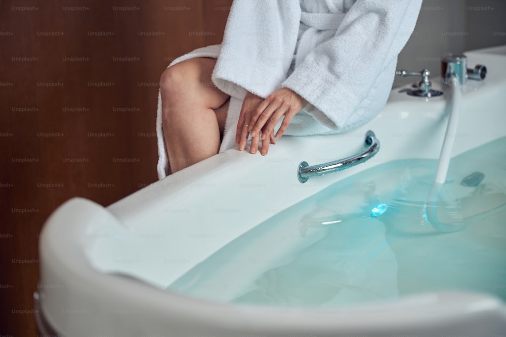 Photo recadrée d’une femme en peignoir éponge assise sur le bord de la baignoire