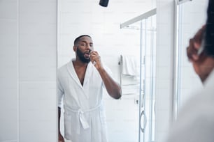 Serious dark-skinned male person wearing bathrobe while doing morning procedures in front of big mirror
