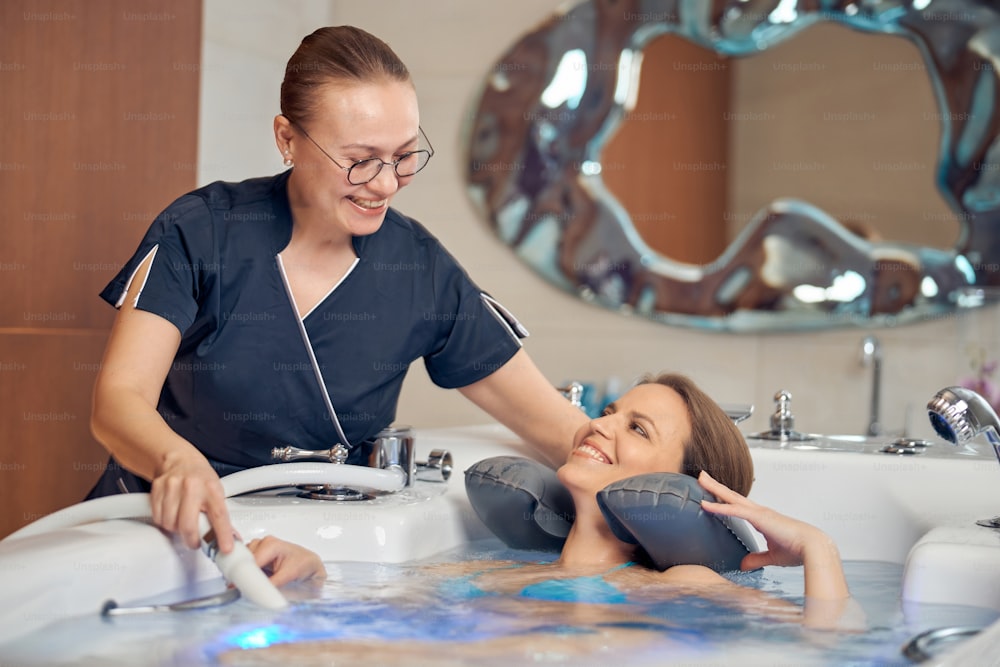 Qualified female therapist in eyeglasses looking at her smiling patient lying in a hydromassage bath