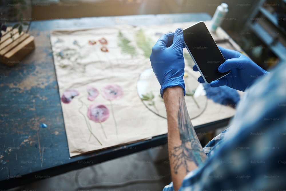 Close up of young man in sterile gloves using smartphone while making herbarium in workshop