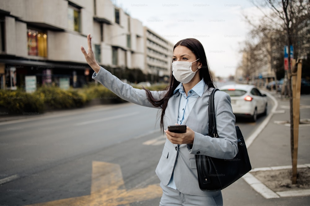 誰もいない街の通りに立ち、危険なインフルエンザやウイルスから身を守るために保護マスクを着用している若くてエレガントなビジネスウーマン。彼女はバス待合所で交通機関を待っています。コロナウイルスまたはCovid-19のコンセプト。
