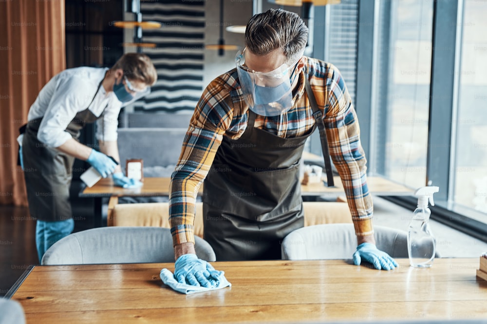 Two male waiters in protective workwear cleaning tables in restaurant