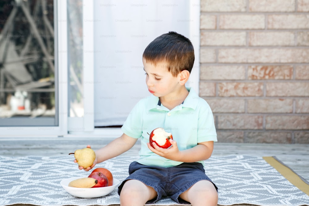 Summer seasonal food. Cute Caucasian preschool boy eating ripe red apple and fruits on backyard. Funny child kid with fresh fruits, apple, banana, pear. Healthy vitamin food for kids children.