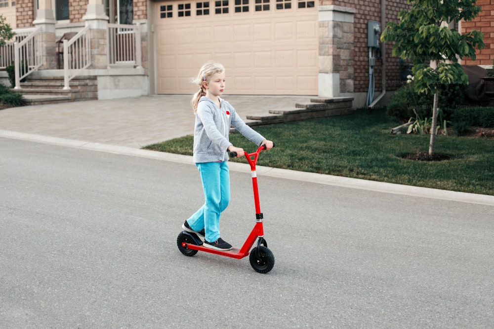 Niña joven que monta scooter rojo en el parque de la calle al aire libre. Diversión de verano actividad deportiva ecológica pasatiempo para niños niños. Auténtico, real, sincero estilo de vida de la infancia.