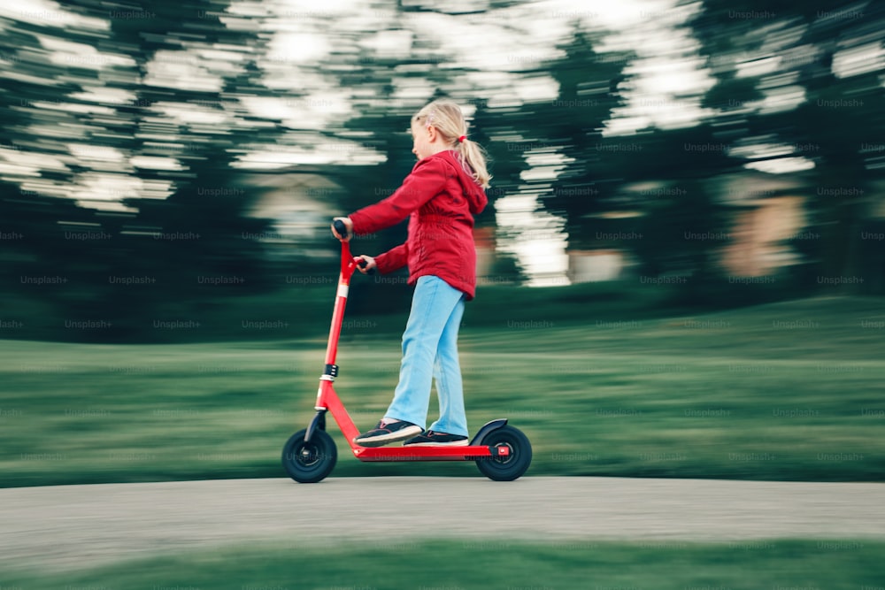 Junges Mädchen in roter Jacke fährt Roller auf Street Road Park im Freien. Sommerspaß Öko-Sport-Aktivität für Kinder Kinder. Action Bewegungsunschärfe. Authentischer, ehrlicher Kindheitslebensstil.