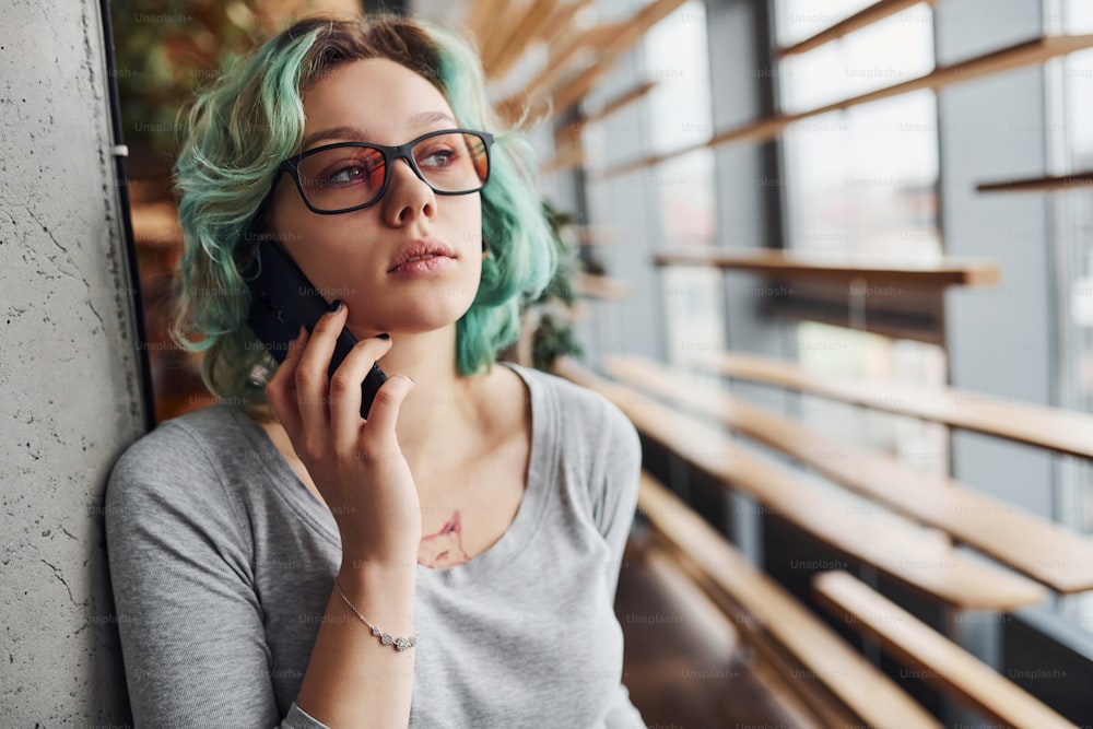 Alternative girl in casual clothes and with green hair standing indoors at daytime and talking by phone.
