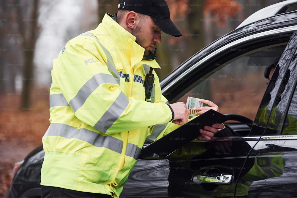 Policial do sexo masculino em uniforme verde recebendo suborno de motorista vehile na estrada.