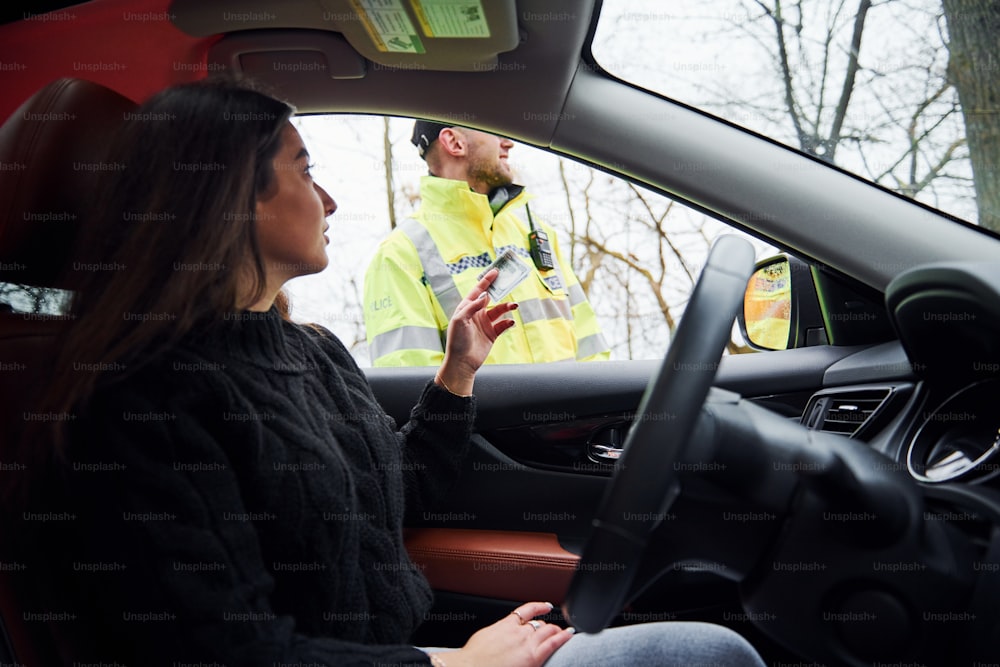 Male police officer in green uniform checking vehicle on the road. Woman trying to give bribe.