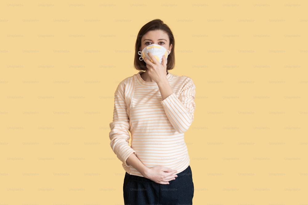 Caucasian pregnant mother is wearing a medical mask posing on a yellow studio wall