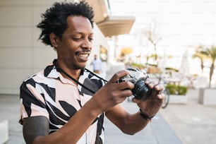 Porträt eines Afro-Touristen, der mit der Kamera fotografiert, während er im Freien auf der Straße spazieren geht. Tourismuskonzept.