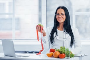 Female nutritionist in white coat holding apple with measuring tape.