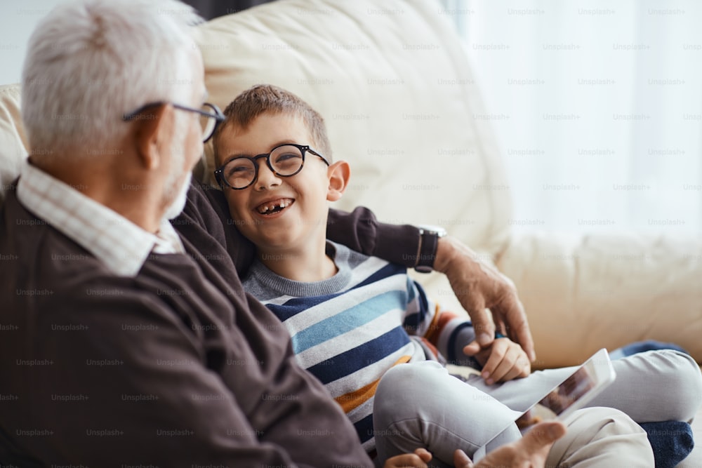 Happy grandson and grandfather talking while sitting embraced on the sofa at home.