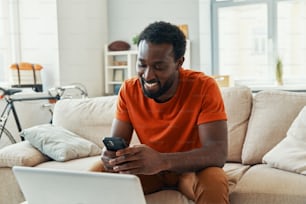 Carefree young African man using smart phone and smiling while spending time at home