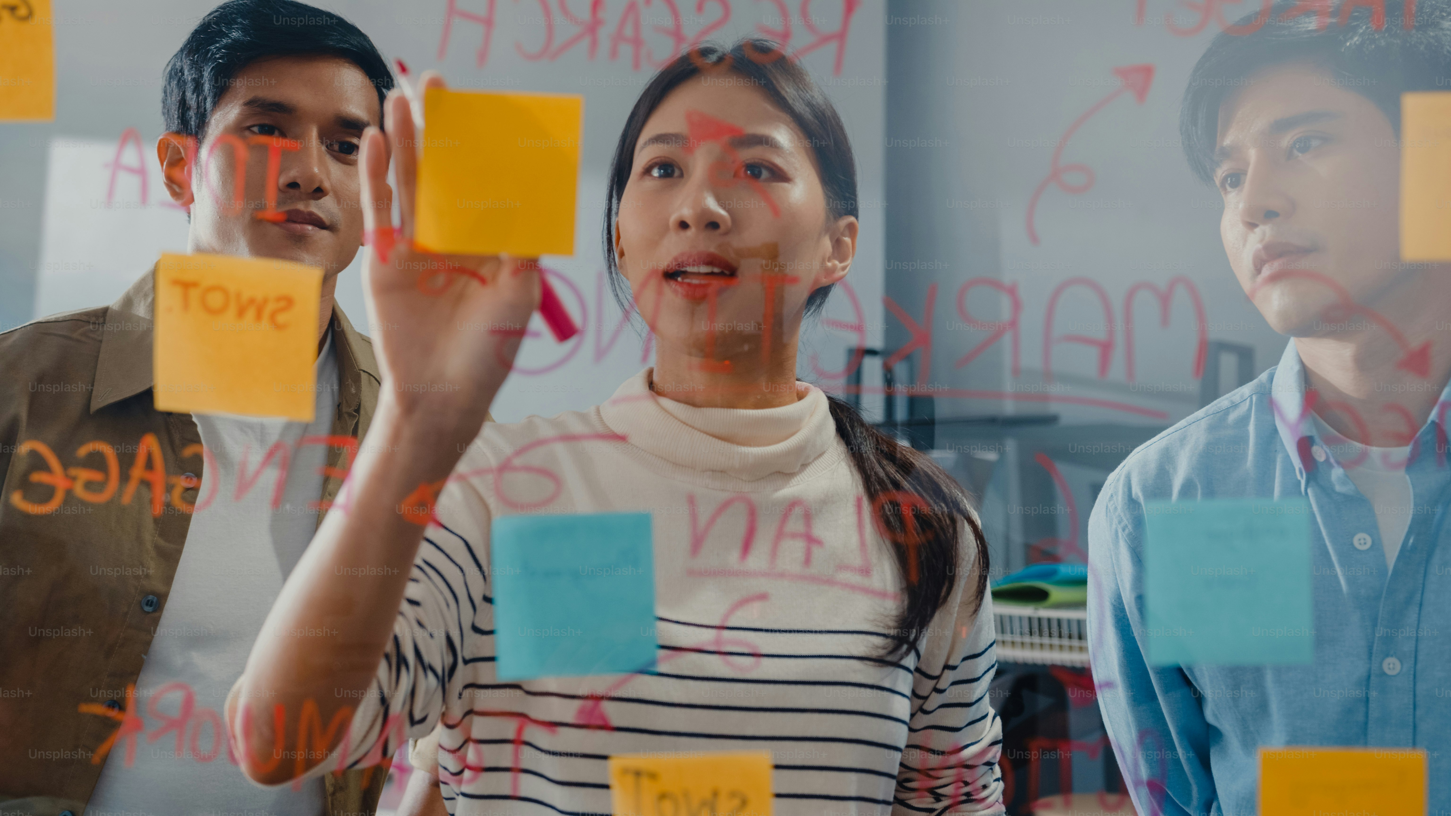 Asia businesspeople stand behind transparent glass wall listen manager pointing progress work and brainstorm meeting and worker post sticky note on wall at office. Business inspiration, Share ideas.