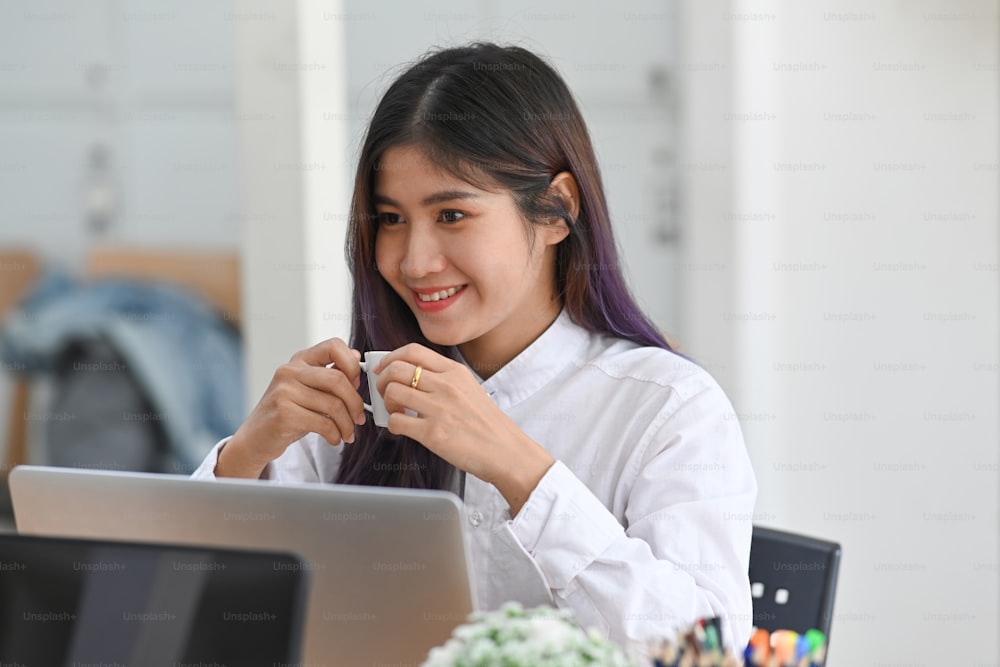 Smiling woman office worker holding coffee cup and sitting at her workplace.