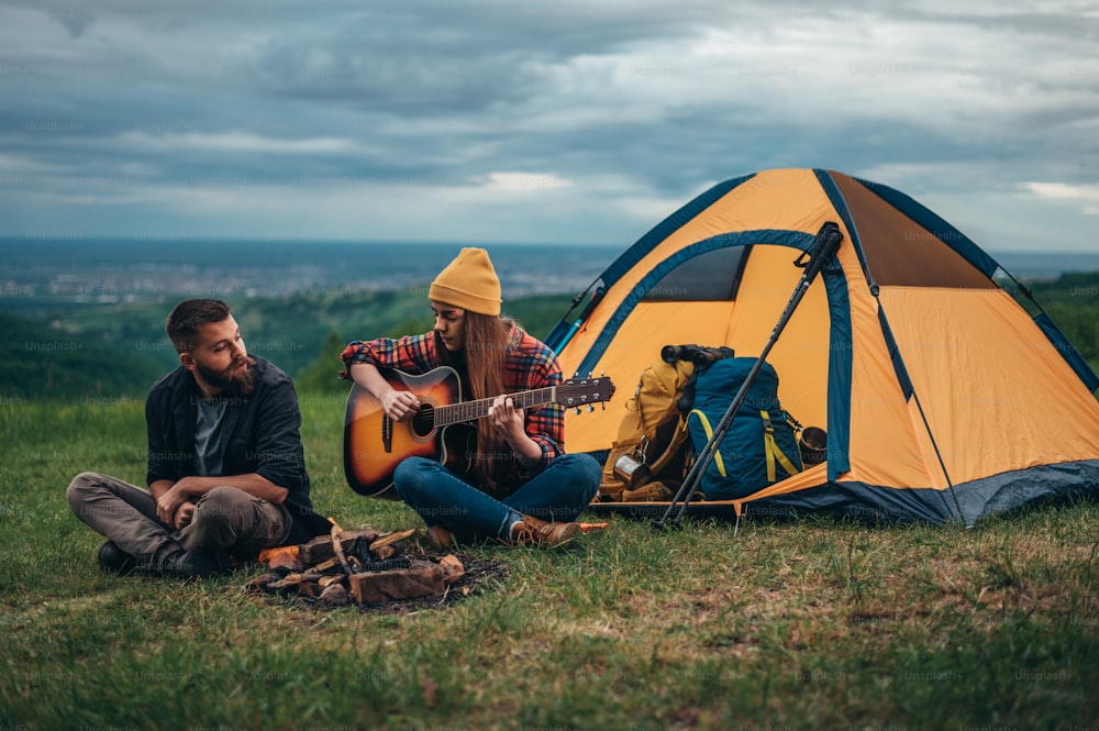 キャンプでテントの近くに座り、楽しみながらギターを弾く若いキャンパーのカップル