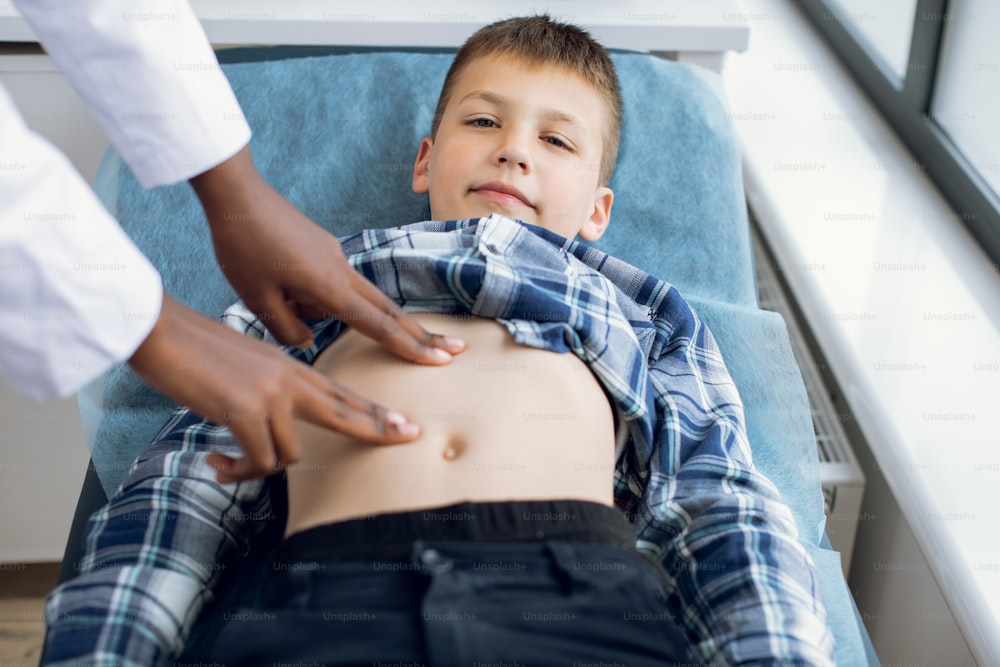 Cute smiling 10-aged kid boy, lying on the couch at modern clinic, visiting his doctor. Female afro pediatrician examining abdomen of child patient using manual palpation