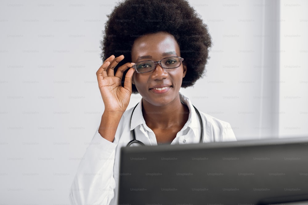 African-American doctor woman, sitting at the table in modern clinic, working on laptop pc, giving remote advice to patients, posing at camera with smile, touching her eyeglasses