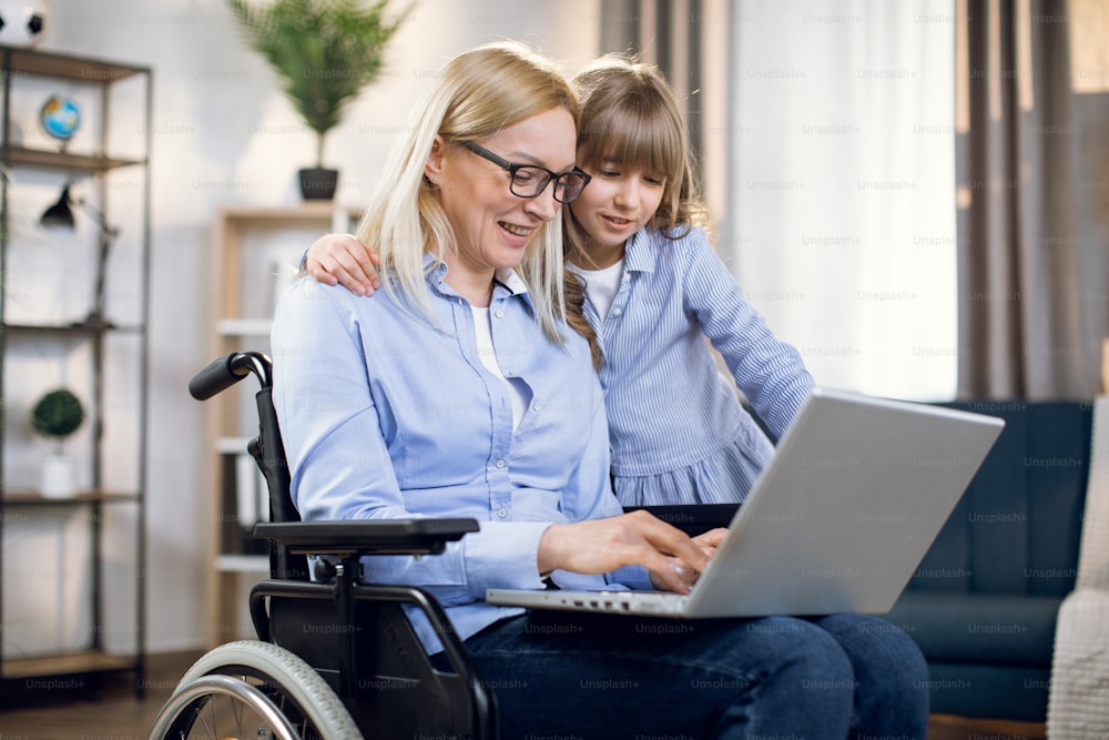 Pretty daughter embracing handicapped mother that sitting in wheelchair with modern laptop on knees. Concept of people, technology and disability.