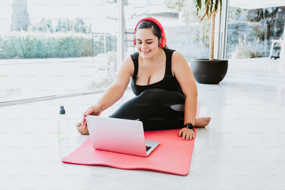 Fille hispanique ronde avec des écouteurs rouges et portant des vêtements de sport prenant un cours de yoga en ligne sur un ordinateur portable à la maison
