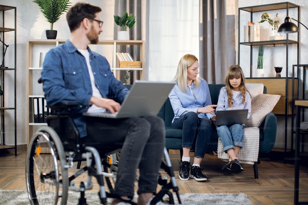 Behinderter bärtiger Mann, der an einem modernen Laptop arbeitet, während seine Frau und seine Tochter zusammen auf dem Sofa sitzen. Remote-Arbeit für behinderte Menschen. Familienkonzept.