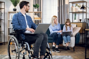 Homme barbu handicapé travaillant sur un ordinateur portable moderne tandis que sa femme et sa fille sont assises ensemble sur un canapé. Travail à distance pour les personnes handicapées. Concept familial.