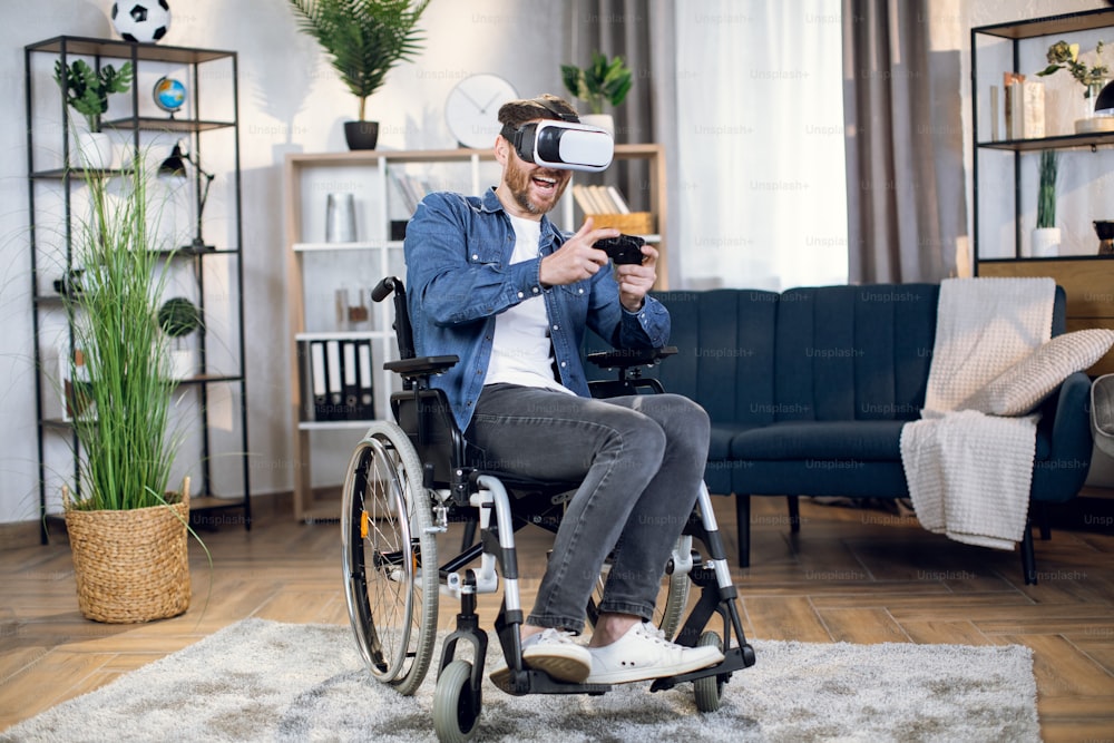 Cheerful handicapped man in virtual glasses playing games using wireless joystick. Entertainment at home for people with special needs. Technology concept.