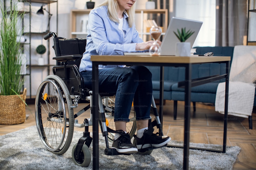 Close up of young woman in casual wear sitting in wheelchair and working on wireless laptop. Disabled female having remote work while staying at home.