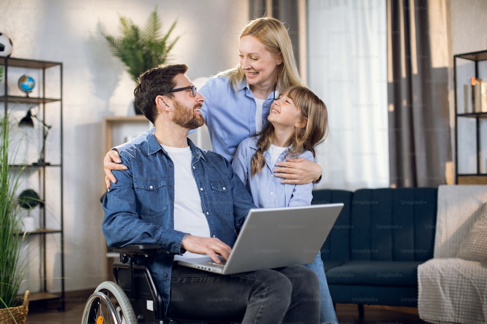 Portrait of bearded man in wheelchair holding laptop on knees while beautiful wife and cute daughter hugging him. Happy caucasian family smiling .