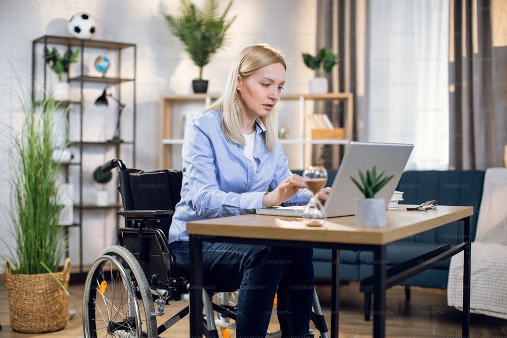 Handicapped woman with blond hair using wireless laptop for remote work from home. Pretty female sitting in wheelchair and typing on keyboard.