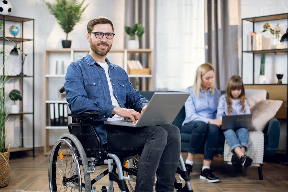 Bearded young man sitting in wheelchair with wireless laptop and smiling on camera. Blur background of beautiful woman and cute child sitting on couch and using computer.
