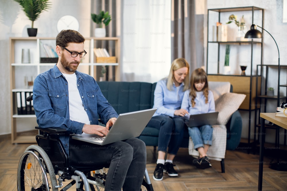장애인 수염을 기른 남자가 현대 노트북으로 작업하는 동안 그의 아내와 딸은 소파에 함께 앉아 있습니다. 장애인을 위한 원격 근무. 가족 개념입니다.