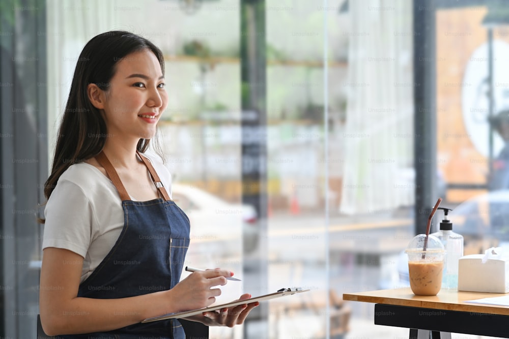 Cameriera asiatica in grembiule che tiene il menu e in piedi nella caffetteria.