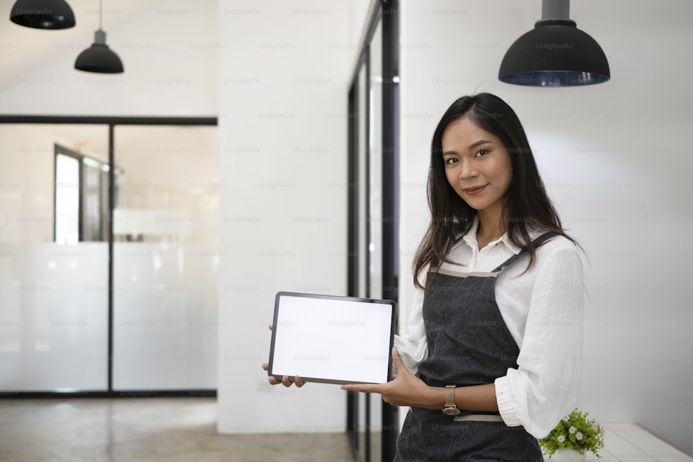 Young woman in apron holding and showing digital tablet with empty screen.