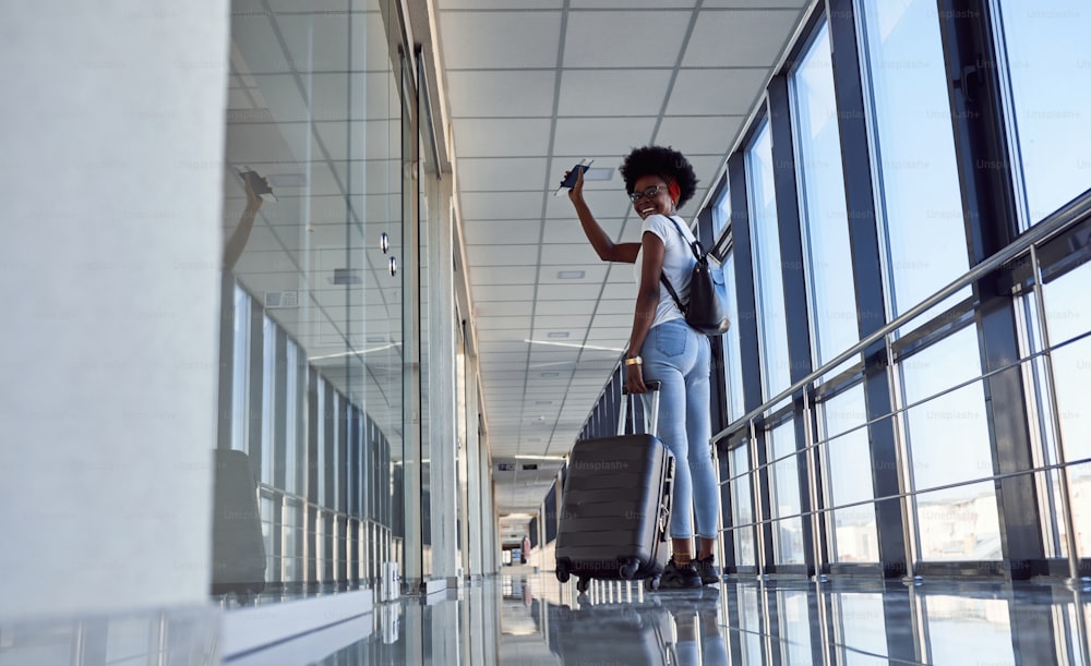 Vista trasera si joven pasajera afroamericana con ropa informal que está en el aeropuerto con equipaje.