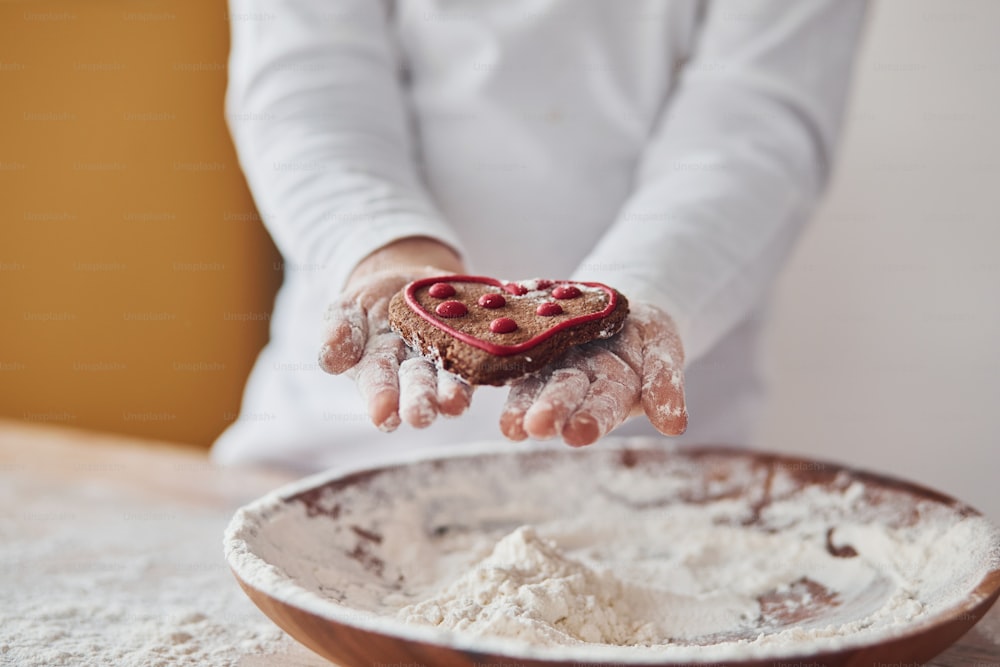 Close up view of little girl's hands that holding sweet cookie on the kitchen with plate of flour.