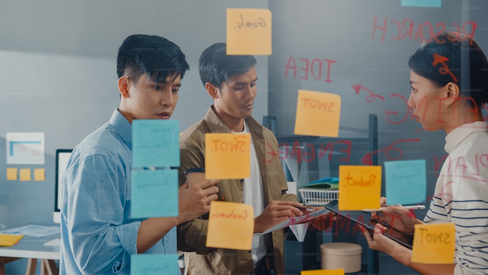 Asia businesspeople stand behind transparent glass wall listen manager pointing progress work and brainstorm meeting and worker post sticky note on wall at office. Business inspiration, Share ideas.