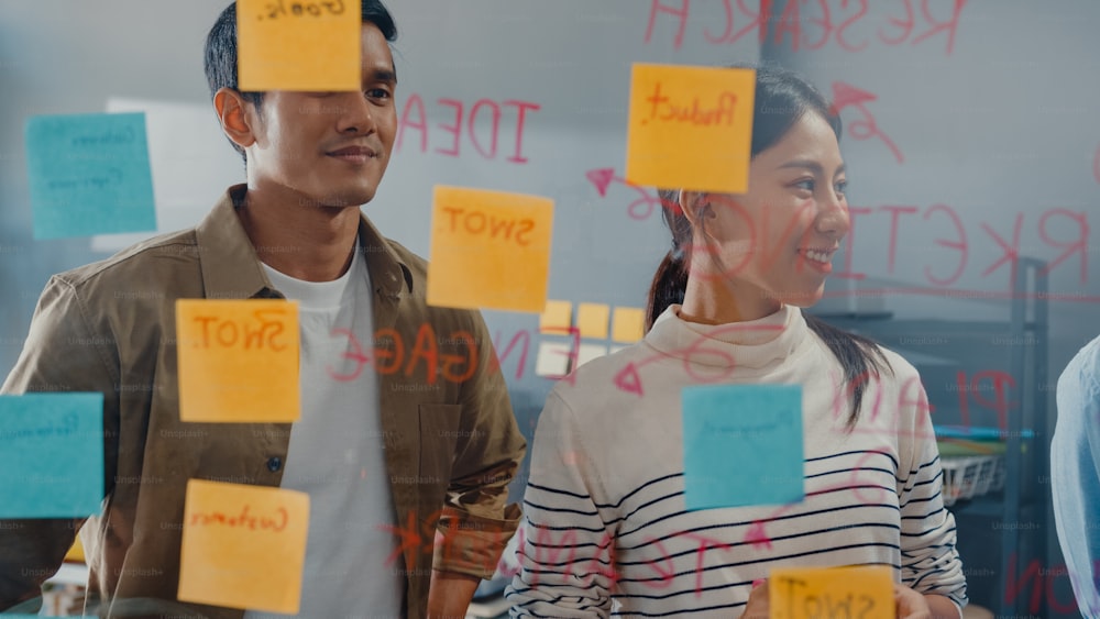 아시아 사업가들은 투명한 유리 벽 뒤에 서서 진행 상황을 가리키는 관리자의 말을 듣고 회의를 브레인스토밍하고 작업자는 사무실 벽에 스티커 메모를 게시합니다. 비즈니스 영감, 아이디어 공유.