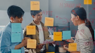 Asia businesspeople stand behind transparent glass wall listen manager pointing progress work and brainstorm meeting and worker post sticky note on wall at office. Business inspiration, Share ideas.