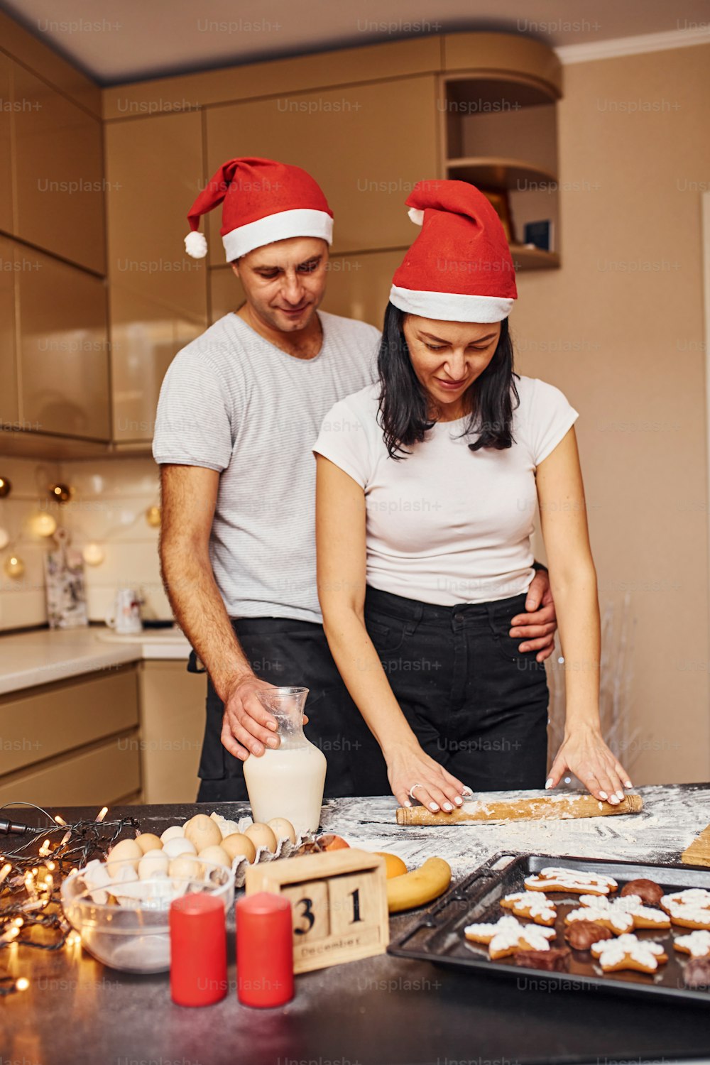 Couple with santa hats standing on the kitchen together indoors.