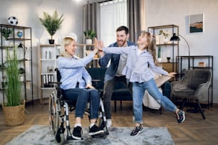 Handicapped woman with blond hair giving high five to handsome husband and pretty daughter at home. Happy parents with child at home. Positive mood of disabled people.