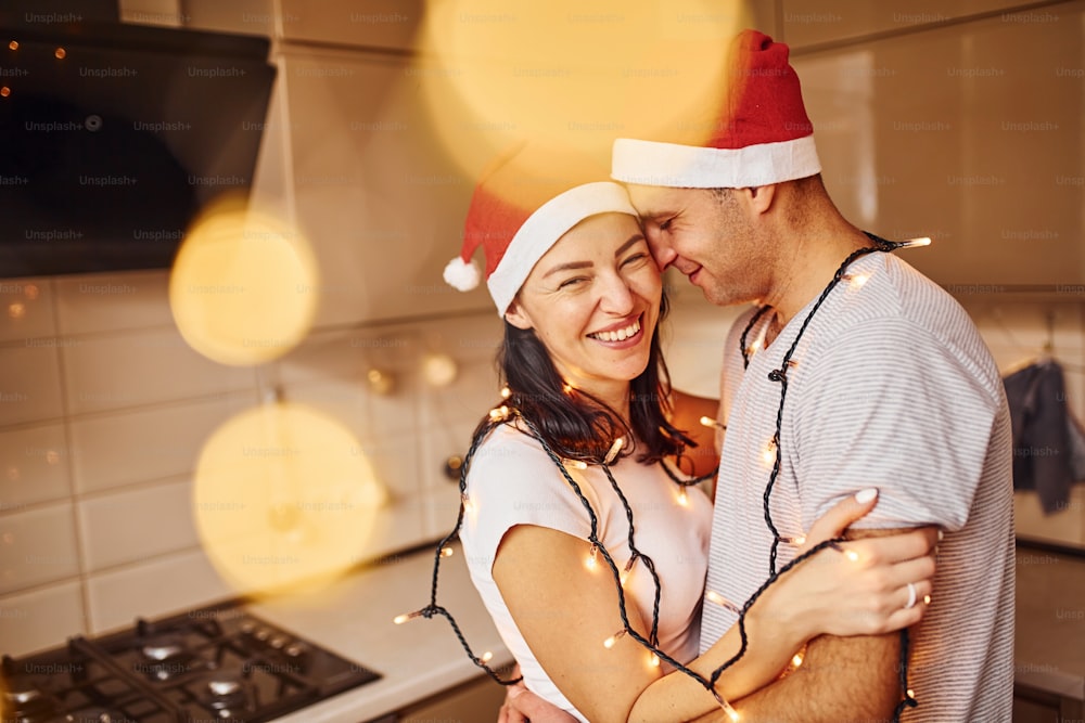 Couple with santa hats standing on the kitchen together indoors.