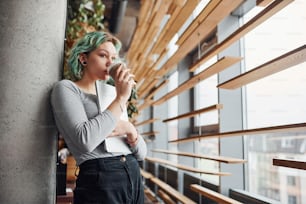 Alternative girl in casual clothes and with green hair standing indoors at daytime with cup of drink in hands.